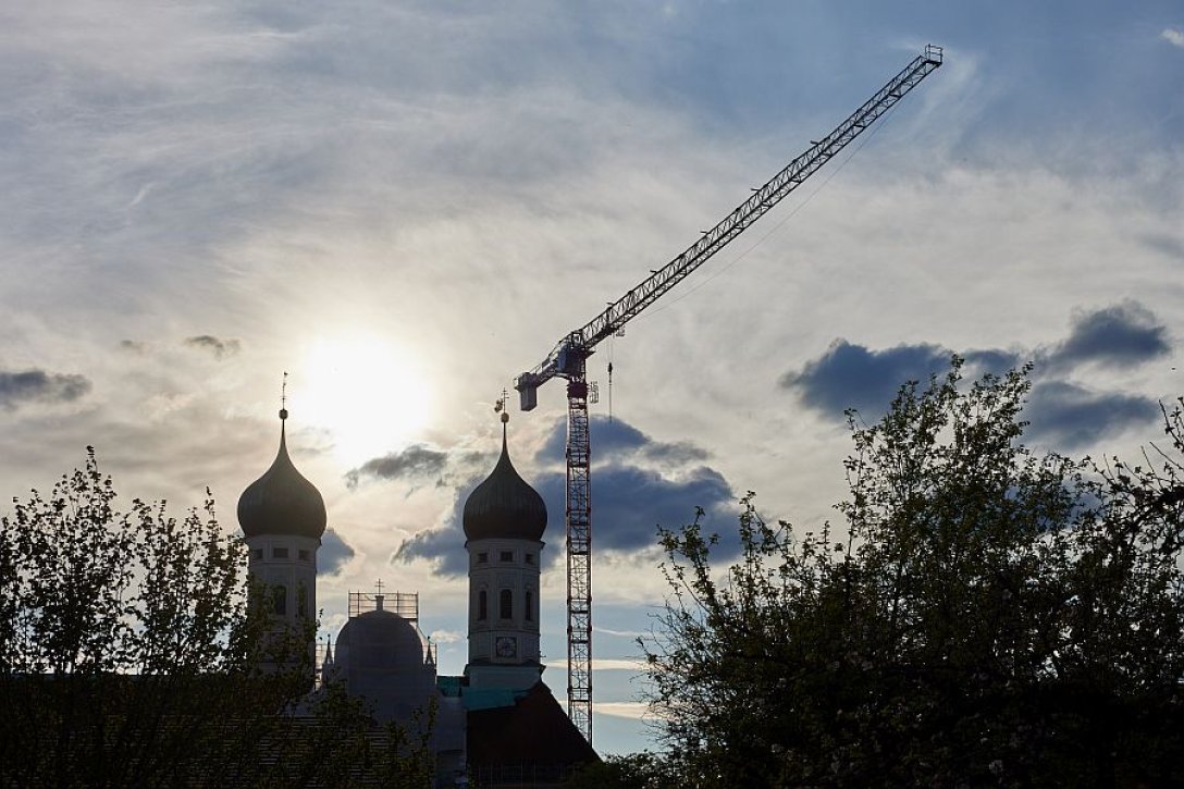 Türme der Basilika St. Benedikt im Kloster Benediktbeuern mit Kran und Gerüsten, dahinter bricht die Sonne durch die dunklen Wolken